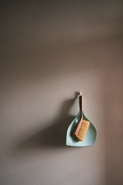 A close-up image of a cleaning tool with a green and blue handle and a flat, rectangular cleaning head, resting on a wooden floor in a residential setting. The cleaning head is covered with a worn-out