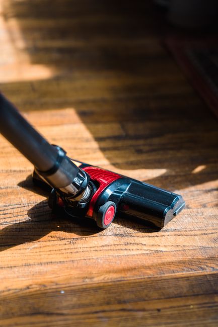 A person wearing grey striped trousers and pink sneakers is using a pink mop to clean a wooden floor in a modern kitchen. A yellow cleaning bucket with a black scrubbing scrubber is placed on the floo