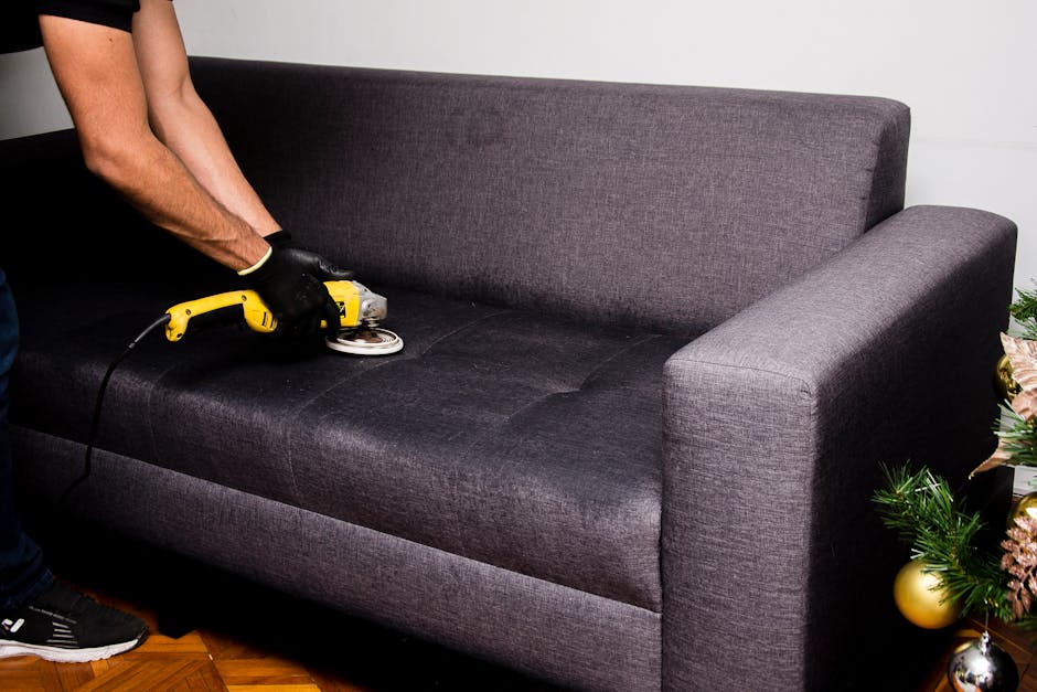 A person wearing black gloves is performing surface cleaning on a dark grey fabric sofa using a handheld upholstery cleaning device with a yellow handle and a round cleaning head. The sofa is situated