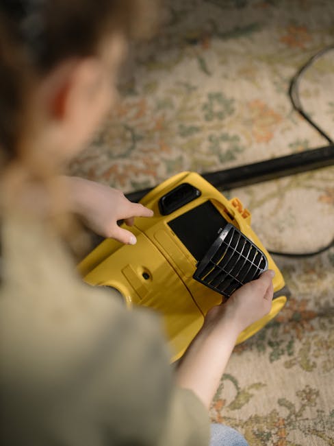 A person wearing black gloves is performing surface cleaning on a dark grey fabric sofa using a handheld upholstery cleaning device with a yellow handle and a round cleaning head. The sofa is situated